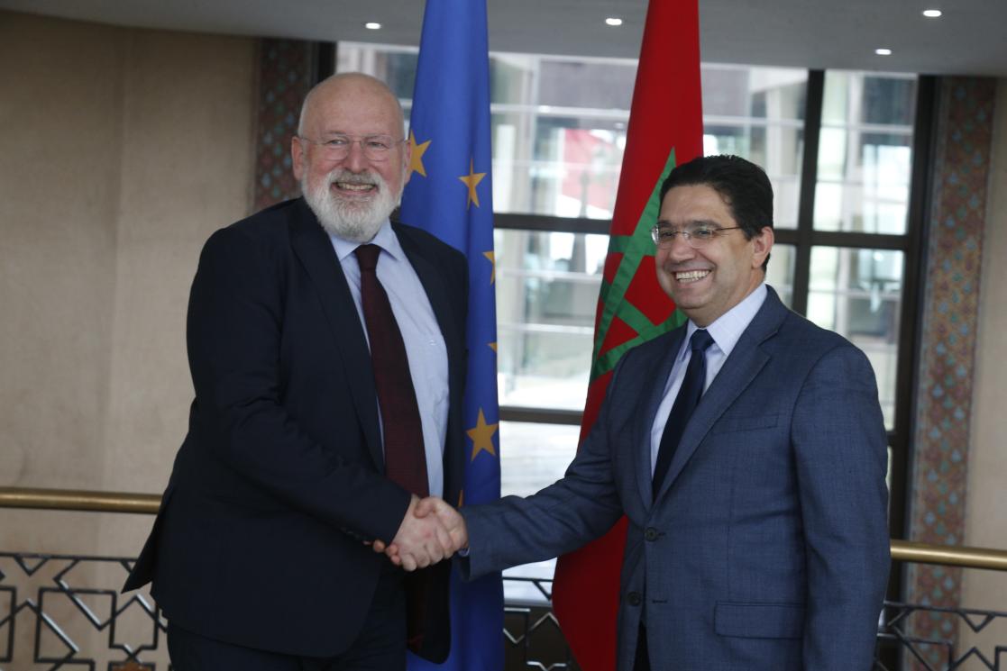 Le Ministre des Affaires étrangères marocain Nasser Bourita et le Vice-Président Exécutif de la Commission européenne Timmermans se serrent la main. Derrière eux le drapeau marocain et le drapeau européen