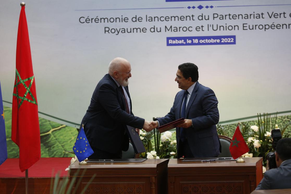 Le ministre des affaires étrangères marocain Bourita et le vice-président exécutif de la commission européenne Timmermans se serrent la main. Ils portent dans leurs mains le document officiel de la signature du Partenariat Vert UE-Maroc et derrière eux le drapeau marocain et européen ainsi qu'une affiche de la cérémonie de lancement.