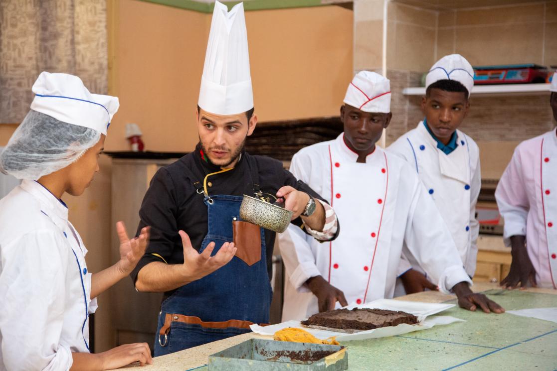 Photo de formation en patisserie à un groupe de jeunes avec une fille, un formateur et deux garçons. Le formateur explique comment faire un gâteau