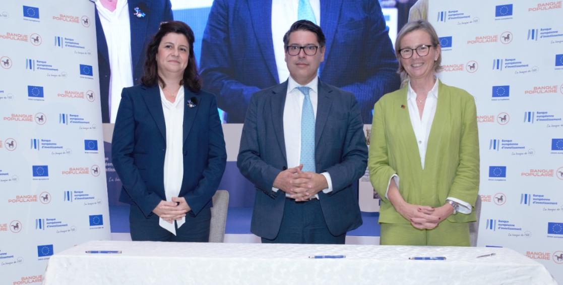 Deux femmes et un homme se tiennent debout les mains jointes, une table devant eux et derrière eux un fond avec les logos de la BEI, de l'UE et de la Banque centrale populaire