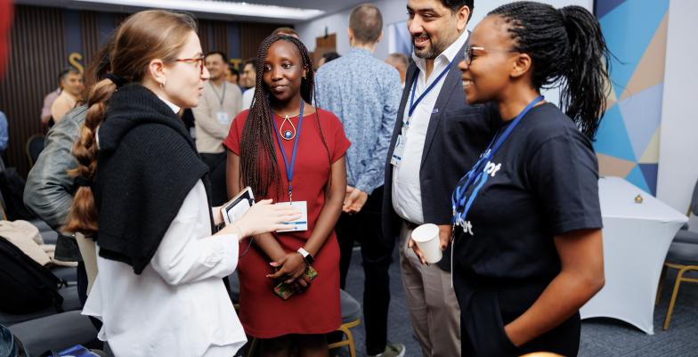 Picture of 4 young media representatives from different countries talking to each other with more people in the background