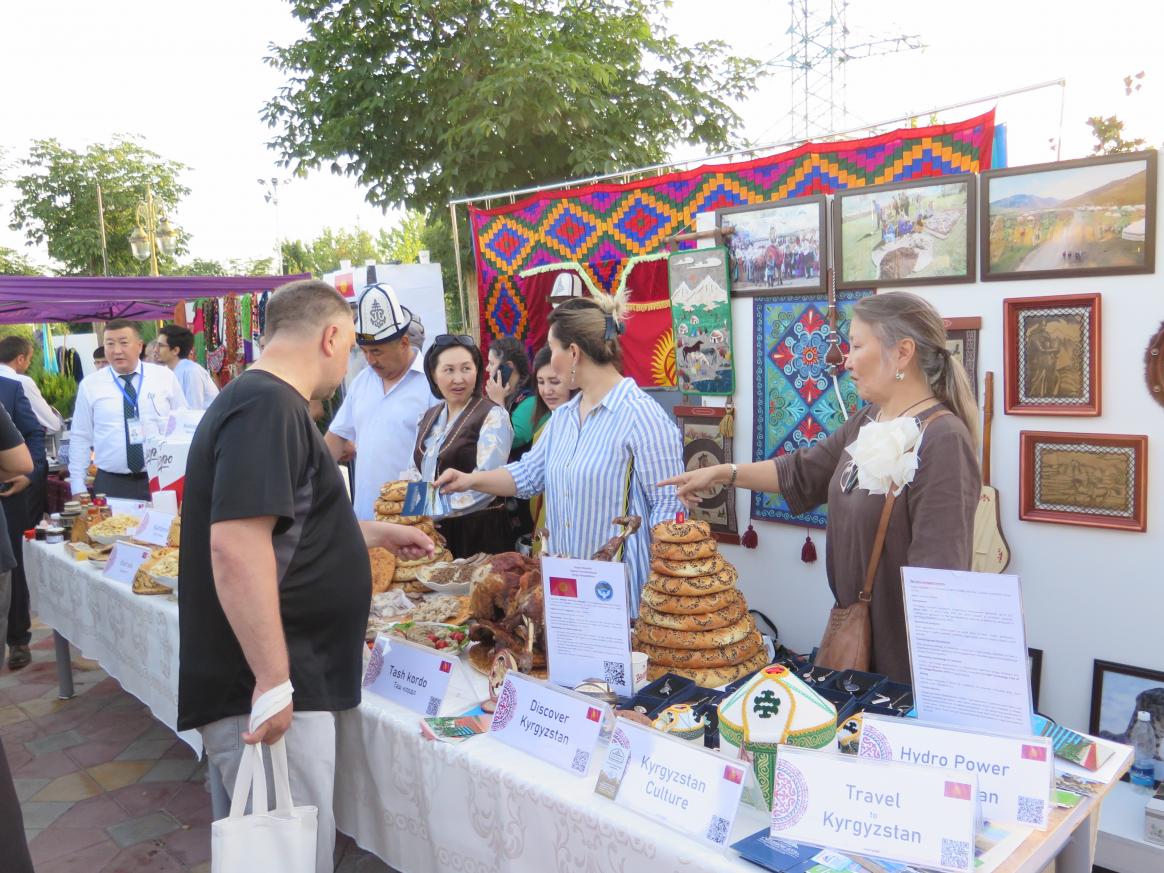 Several people behind a stand filled with different breads and nametags. Behind them hang pictures and fabrics. One person on the other side of the stand pointing to one bread.