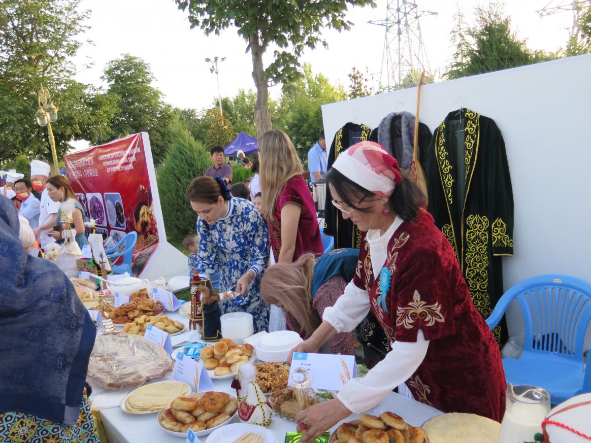 Four people behind a stand filled with different foods with tags next to them. Behind the people are three coats hanged on the wall.