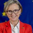 une femme habillée en rouge avec des lunettes, posant souriante avec derrière un fond bleu ou on devine les étoiles du dapeau de l'UE
