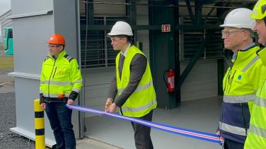 Three people wearing bright jackets. One person in the middle cutting a blue and red ribbon.