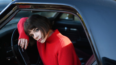 A woman in a red turtleneck sweater leans out the window of a car.