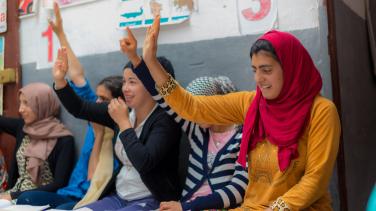 cinq jeunes filles avec les mains levées dans une salle de classe avec derrière un mur avec des dessins scolaires