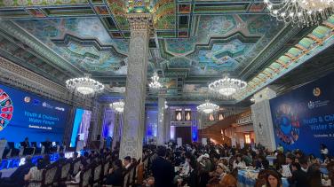Big hall decorated ceiling and columns, chandeliers in the ceiling. People sitting in rows facing left, where behind a stage reads Youth & Children's Water Forum.