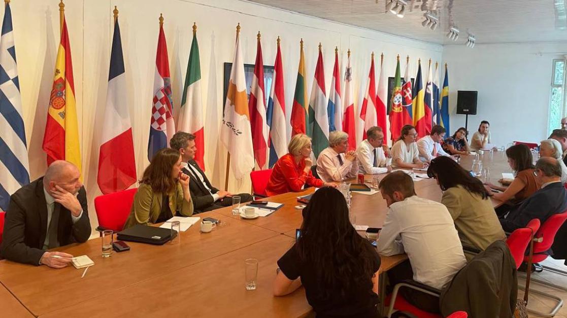 A group of people sitting around a large table with flags in the room all around. 