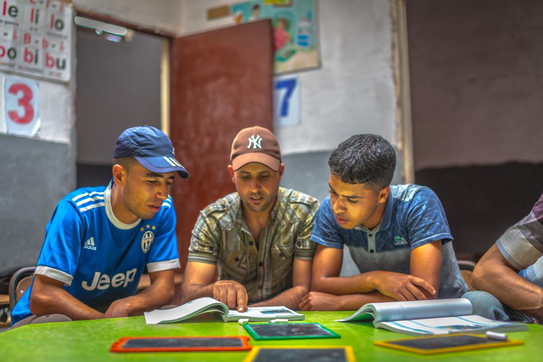 Trois jeunes hommes dans une salle de classe en train de lire dans un cahier déposé sur une tableau avec des ardoises et derrière eux une porte et des murs avec des dessins