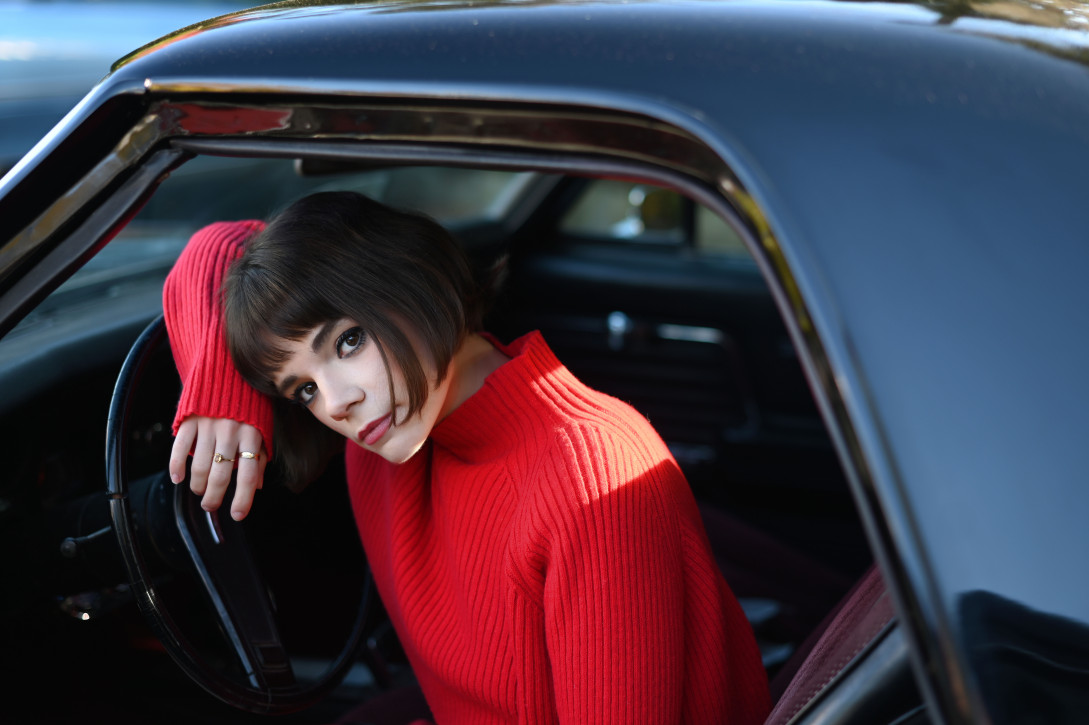 A woman in a red turtleneck sweater leans out the window of a car.