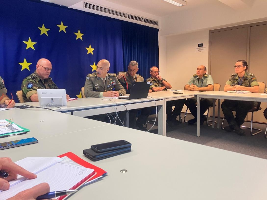 A meeting of senior military personnel in an office with a notebook in the foreground. There is an EU flag on the wall. 
