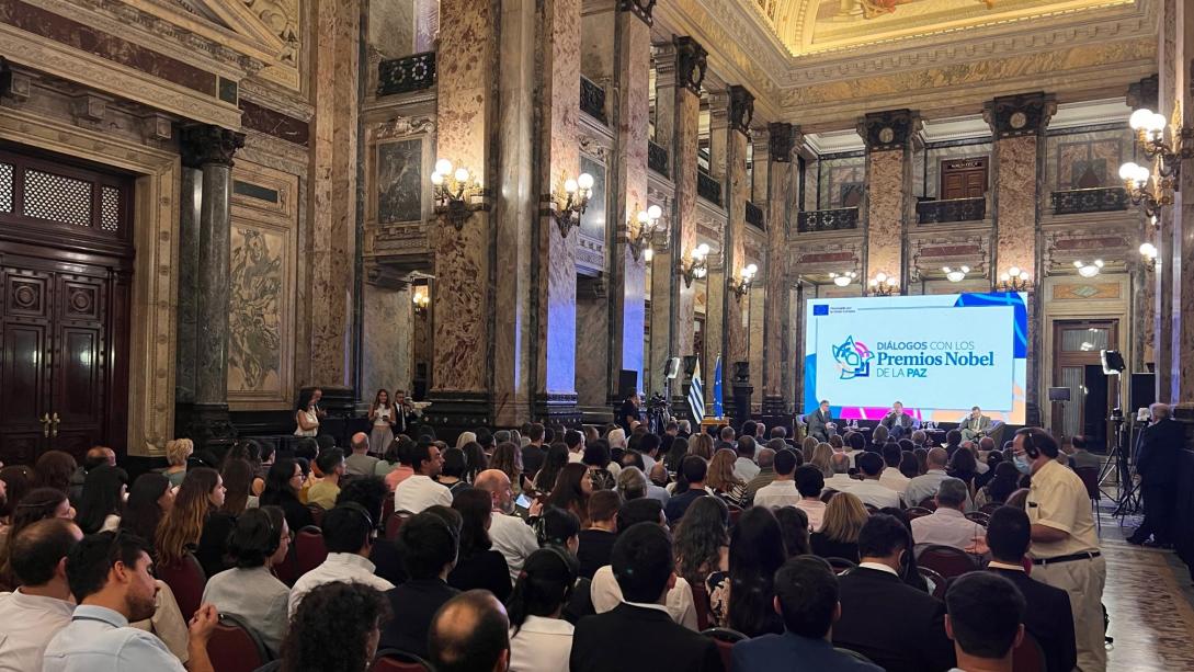 An audience in a decorative room listens to speeches in the background