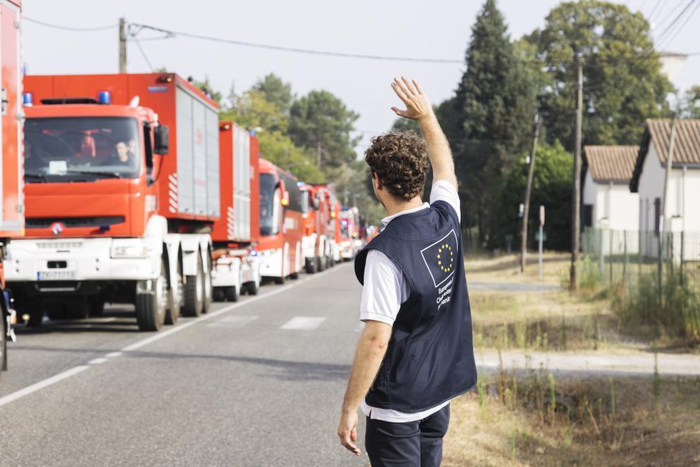 Man wearing a vest with the EU logo printed on the it, standing and waving to firefighter trucks