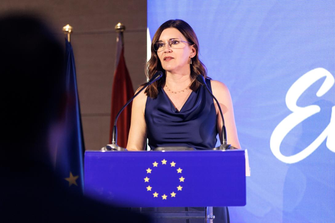 Oana Cristina Popa standing on stage in a blue dress. Behind her is a screen and two flags