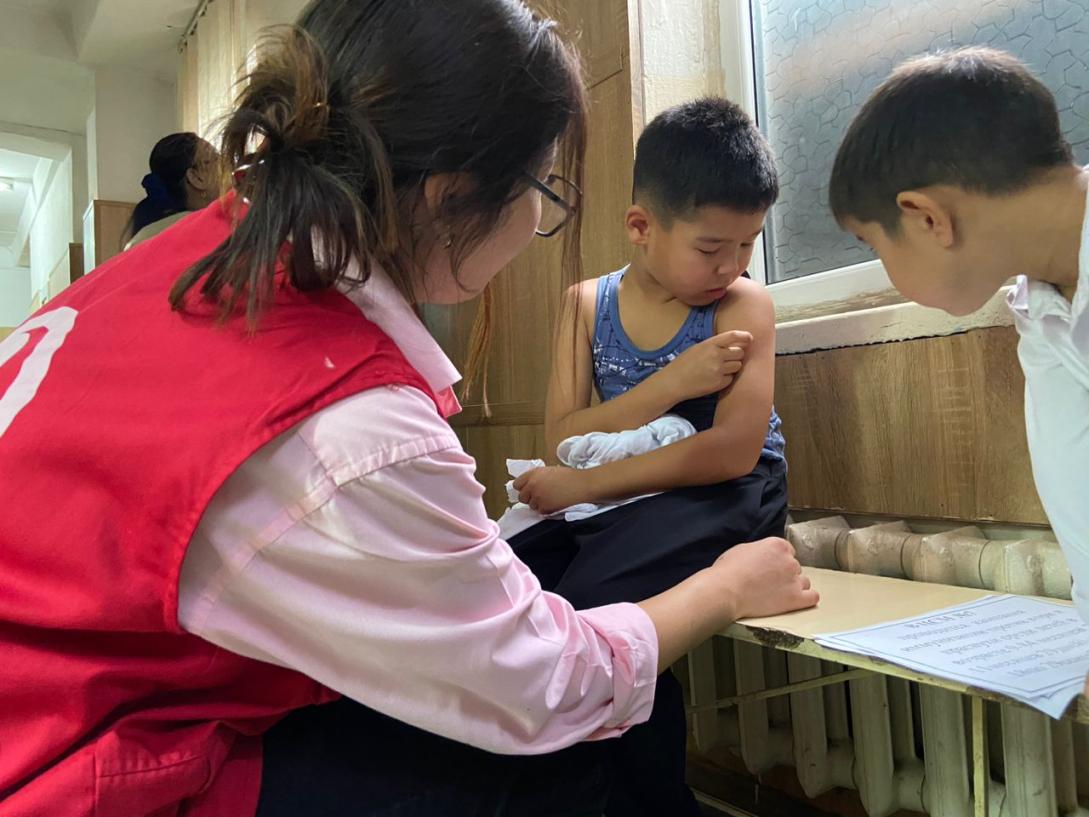A child receives medical attention from an adult in a red vest while another child observes.