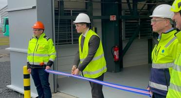 Three people wearing bright jackets. One person in the middle cutting a blue and red ribbon. 