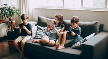 Three children on a gray sofa engrossed in digital devices, indoors.