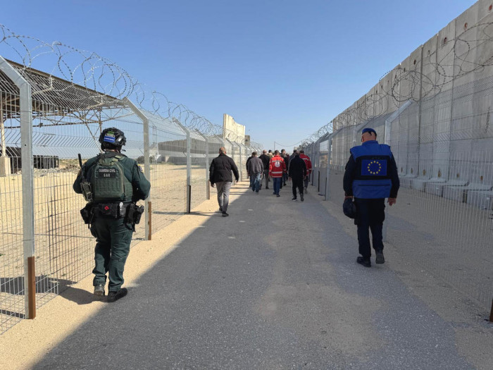 Exterior, uniformed people watch civilians at the border crossing. 