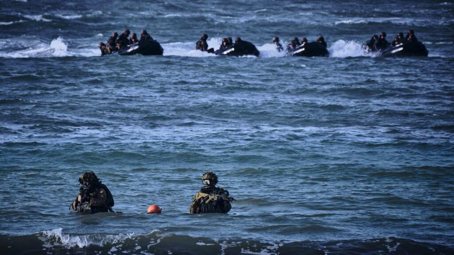 Background, dozens of combat personnel approach the shore in inflatables on mission, two combat divers in the foreground.