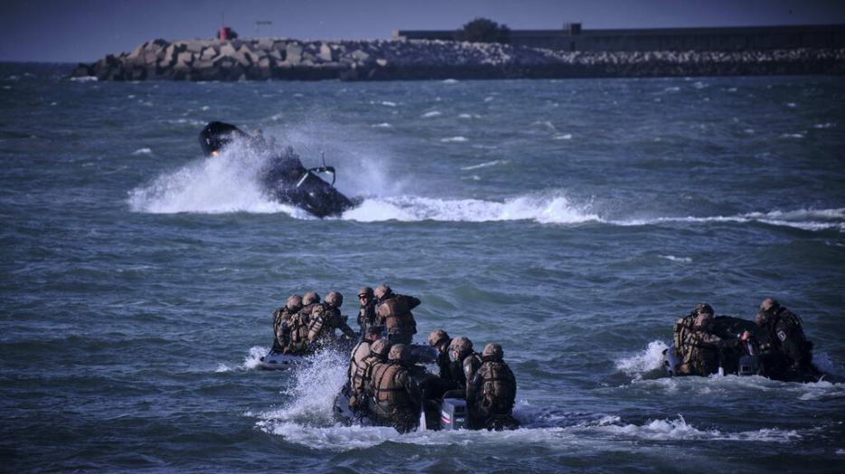 Dozens of combat personnel manouvre close to shoreline in boats during amphibious exercise. 