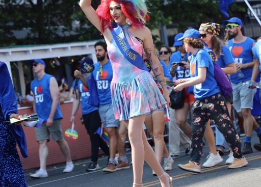 A drag queen in a rainbow wig waves at parade onlookers.