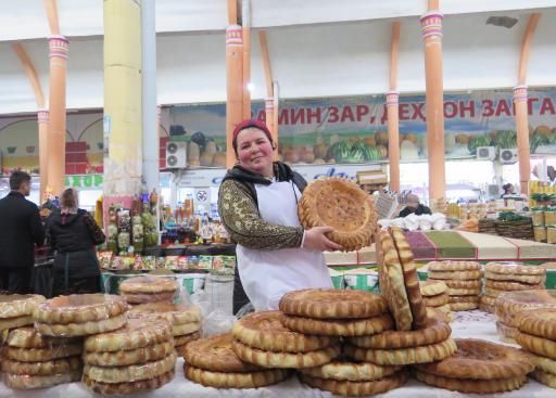 Person holding a round bread while posing and smiling for the camera behind a counter stacked with more bread.