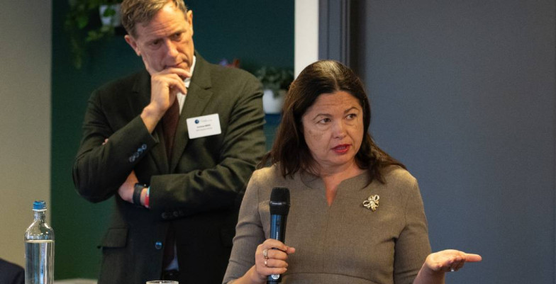 A speaker in foreground with the chairperson in background listening closely.