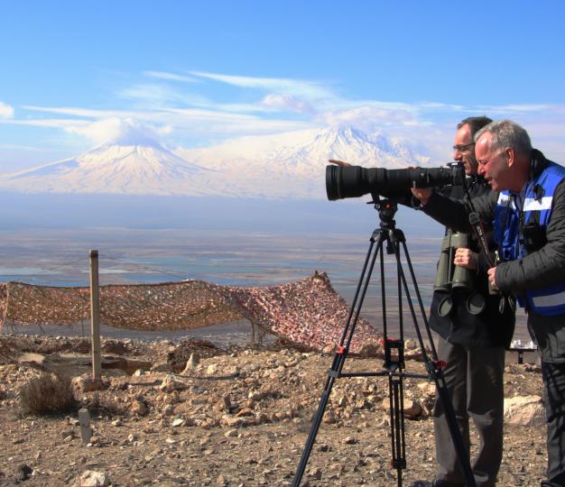 Two men with optical instruments look out over a mountainous landscape.
