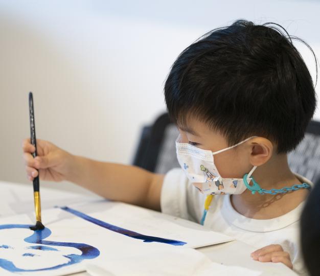 A little boy with black hair paints with blue paint at a table.