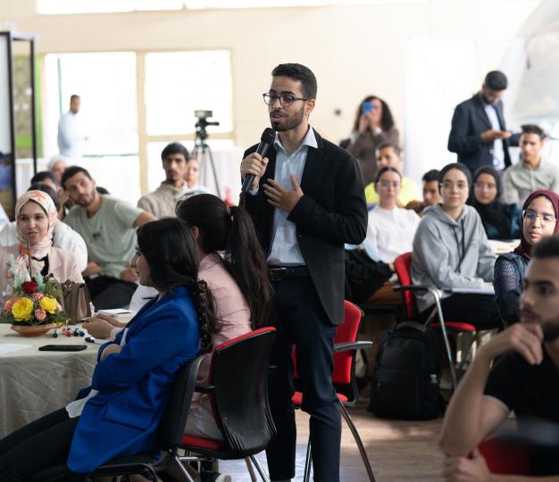 Jeune homme qui porte des lunettes et parle dans un micro debout devant un groupe de jeunes assis dans un format tables rondes