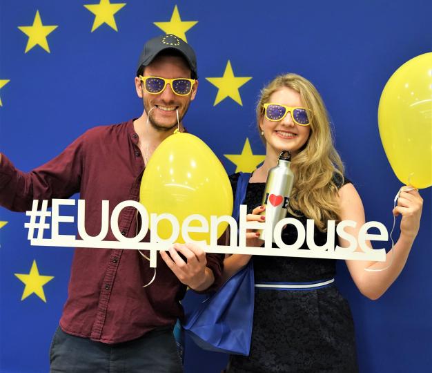 A man and a woman wearing EU sunglasses hold yellow balloons and an EU Open House sign