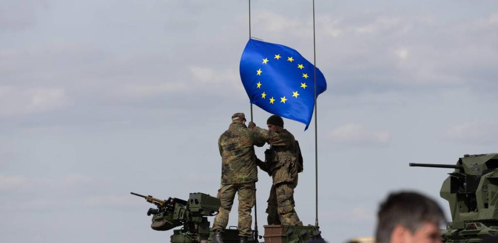 Two soldiers on an armoured vehicle hoisting the EU flag.